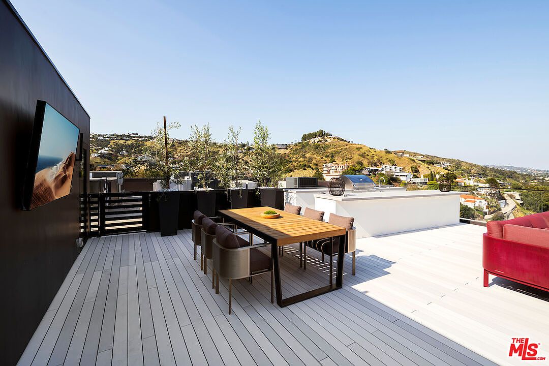 This expansive rooftop deck features light-toned composite decking and a modern dining set, perfect for outdoor entertaining. The space includes a built-in outdoor kitchen area with a grill and a wall-mounted television, all set against a backdrop of scenic hillside views. The clean, contemporary design creates an inviting atmosphere for enjoying the surrounding landscape.