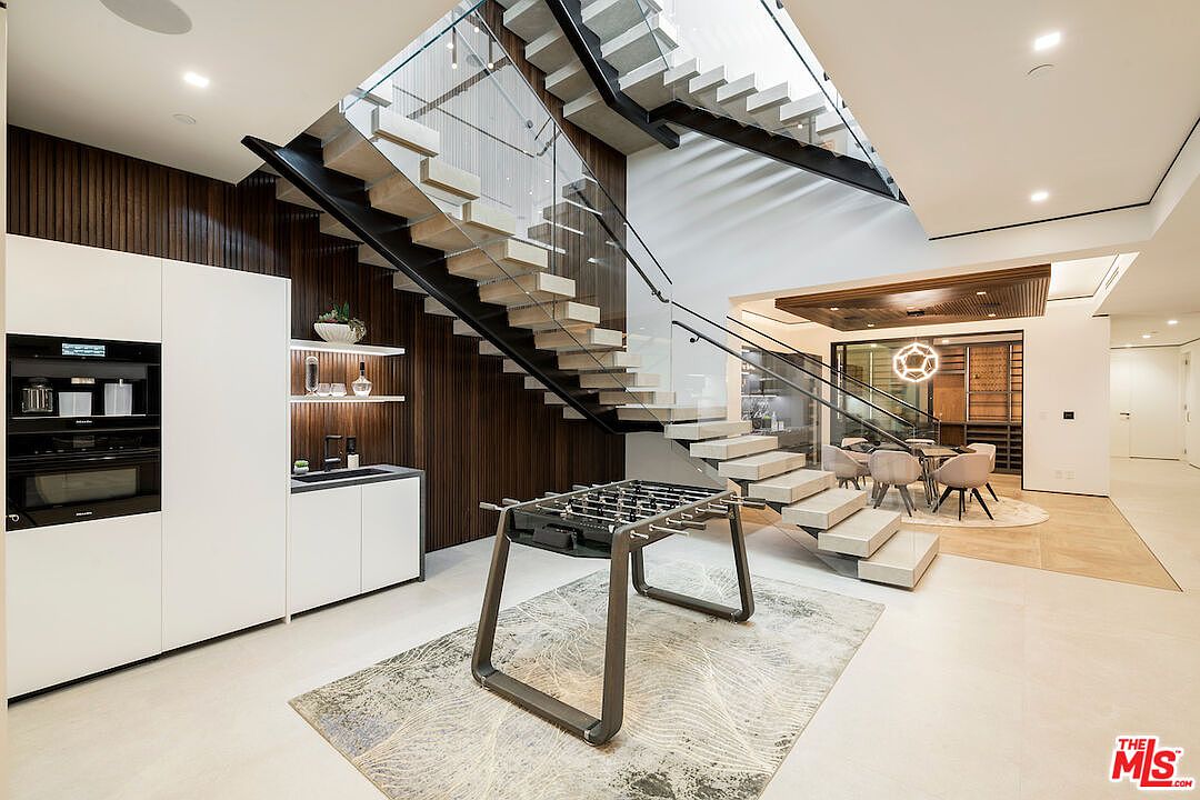 This modern interior space features a striking floating staircase with light wood treads and glass railings, serving as a central architectural focal point. Below the stairs, a foosball table sits on a patterned area rug, while a sleek wet bar with white cabinetry and dark wood paneling is integrated into the wall. The perspective looks toward a dining area in the background, emphasizing the open-concept layout and contemporary design aesthetic.