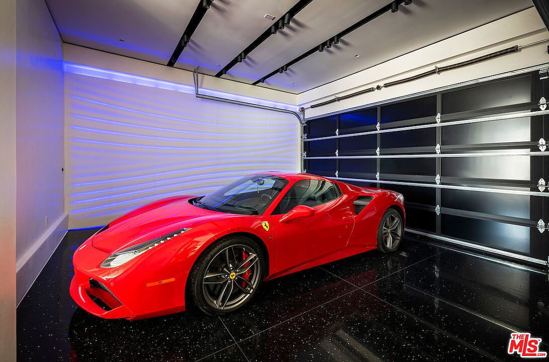 This high-end, modern garage features a sleek, polished black floor with subtle metallic flecks, providing a dramatic backdrop for a bright red Ferrari. The space is illuminated by recessed track lighting and a vibrant blue LED accent strip along the textured, wavy wall. A large, multi-panel black garage door with horizontal glass inserts completes the sophisticated, showroom-like aesthetic.