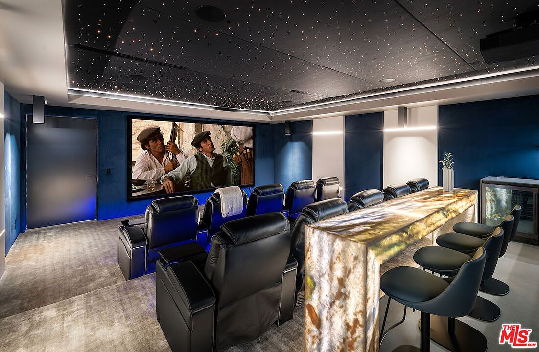 This luxurious home theater features a tiered seating arrangement with plush black leather recliners facing a large projection screen. The room is accented by a dramatic starlit ceiling and a backlit stone bar counter with modern stools, creating an immersive cinematic atmosphere. The space is finished with deep blue wall panels and soft carpeting, providing a sophisticated and comfortable environment for entertainment.