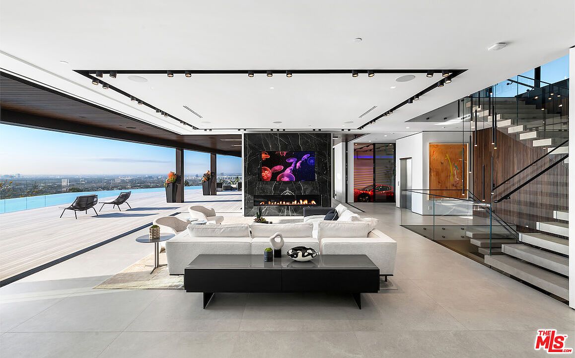 This expansive, modern living room features an open-concept layout that seamlessly transitions to an outdoor deck through large glass walls. A central dark stone-clad chimney breast serves as a focal point, housing a television and a linear fireplace. The space is furnished with a large white sectional sofa, a sleek black coffee table, and a low-profile console, all set against a backdrop of neutral tile flooring and a floating staircase with glass railings.