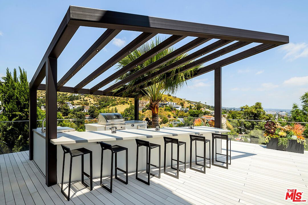 This elevated outdoor deck features a modern, minimalist bar area sheltered by a sleek, dark-toned pergola. The space is equipped with a built-in grill and four contemporary bar stools, offering a panoramic view of the surrounding hillside landscape. The clean lines and open-air design create a sophisticated setting for outdoor entertaining.