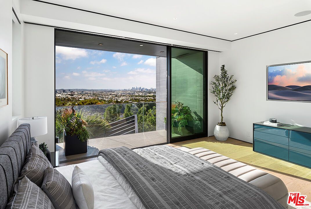 This modern primary bedroom features a plush, dark-toned upholstered bed and a sleek, teal-colored dresser. A large sliding glass door opens onto a private balcony, providing an expansive, unobstructed view of the city skyline. The room is bright and airy, with light wood flooring and a minimalist aesthetic that emphasizes the indoor-outdoor connection.