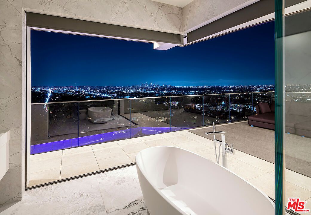 This luxurious primary bathroom features a sleek, freestanding white soaking tub positioned to overlook a panoramic city skyline at night. The space is defined by expansive floor-to-ceiling glass walls that seamlessly connect the interior to an outdoor balcony, creating an open and airy atmosphere. The room is finished with light-toned stone flooring and modern fixtures, emphasizing a sophisticated, high-end contemporary design.