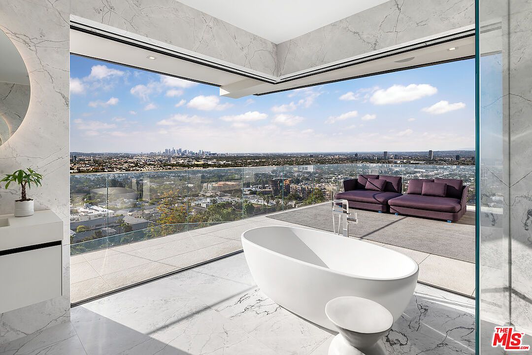 This luxurious primary bathroom features a freestanding soaking tub positioned to take advantage of expansive, panoramic city views through large, corner-wrapping glass walls. The space is finished with light-toned, veined stone surfaces and includes a private balcony area furnished with plush purple lounge seating. The overall aesthetic is ultra-modern and sophisticated, emphasizing a seamless transition between indoor comfort and the outdoor cityscape.