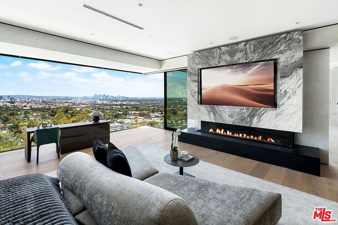 This sophisticated living room features a striking stone-clad chimney breast that serves as a focal point, housing a wide linear fireplace and a wall-mounted television. The space is defined by expansive glass walls that provide panoramic city views, complemented by light wood flooring and a plush, neutral-toned sectional sofa. A minimalist desk area is positioned near the window, creating a seamless blend of luxury living and functional workspace.