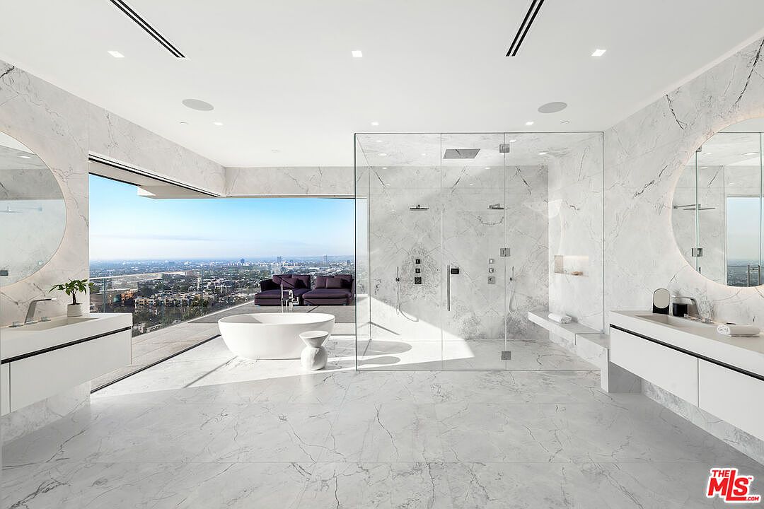 This luxurious primary bathroom features a minimalist, open-concept design with extensive use of light-toned stone surfaces throughout. A freestanding soaking tub is positioned in front of floor-to-ceiling glass walls that provide an expansive, panoramic view of the city skyline, while a large, glass-enclosed walk-in shower sits centrally in the room. Dual floating vanities with circular mirrors flank the space, creating a symmetrical and sophisticated aesthetic.
