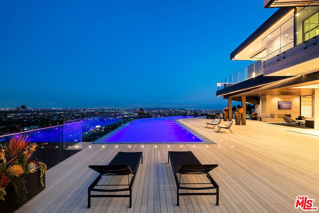 This stunning twilight view captures a modern infinity-edge pool set against a sprawling deck that overlooks a vast city skyline. The deck features sleek lounge chairs and integrated lighting, creating a sophisticated outdoor living space that seamlessly blends with the horizon. The architecture showcases clean lines and expansive glass railings, emphasizing a luxurious and contemporary aesthetic.