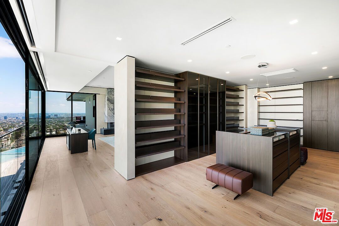 This expansive, modern walk-in closet features a central island with drawers and a leather-topped bench, surrounded by dark wood shelving and glass-front cabinetry. The space is illuminated by recessed lighting and a contemporary pendant fixture, while floor-to-ceiling glass walls provide a panoramic view of the city skyline. The open-concept design seamlessly transitions into a bright office area, emphasizing a luxurious and airy atmosphere.
