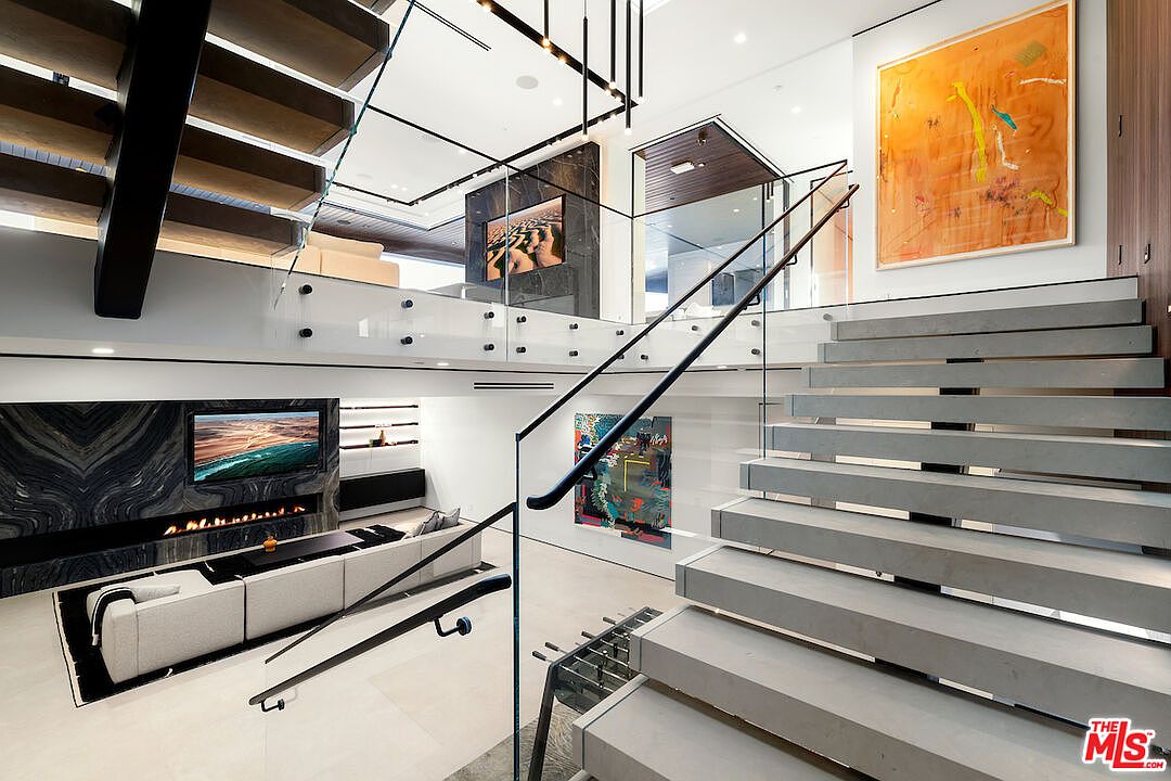 This modern, multi-level interior space features a prominent floating staircase with thick, light-toned treads and a glass railing. The open-concept design connects a lower-level living area, characterized by a dark stone-clad chimney breast and a large television, to an upper landing. The aesthetic is sleek and contemporary, highlighted by minimalist lighting fixtures and vibrant abstract artwork on the walls.
