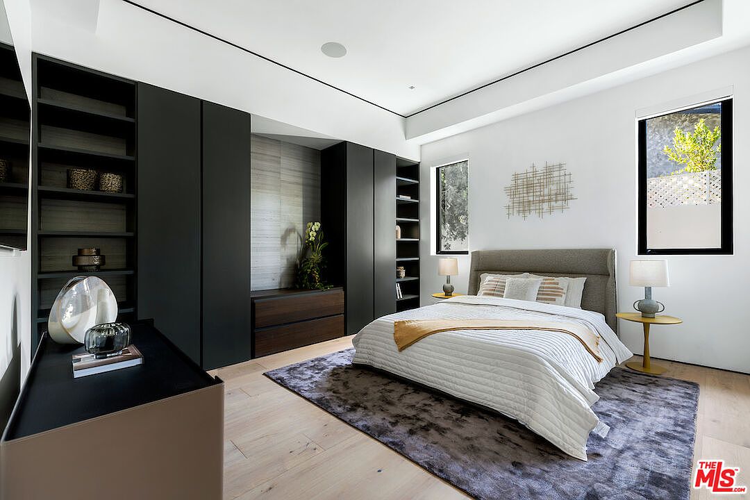 This contemporary bedroom features a neutral color palette with a large, upholstered bed as the focal point, set atop a textured area rug. A wall of dark, built-in cabinetry provides ample storage and display shelving, while light wood flooring adds warmth to the space. The room is illuminated by natural light from two windows and features a minimalist aesthetic with clean lines and modern decor.