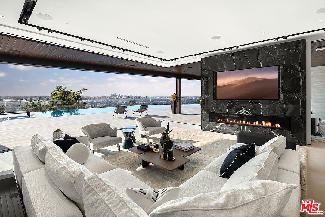 This expansive, modern living room features an open-concept design that seamlessly transitions to an outdoor deck through large, retractable glass walls. The space is anchored by a striking, dark stone-clad chimney breast housing a wide, linear fireplace and a mounted television. A plush, light-colored sectional sofa and contemporary armchairs are arranged on a textured rug, creating a sophisticated lounge area overlooking a swimming pool and a panoramic city skyline.