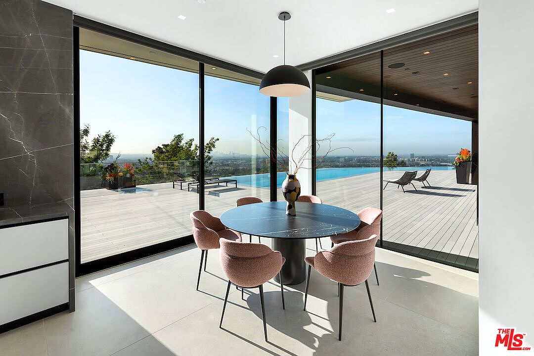 This contemporary dining area features a round, dark stone-topped table surrounded by five upholstered pink chairs. The space is defined by expansive floor-to-ceiling glass walls that provide a seamless transition to an outdoor deck and an infinity-edge pool overlooking a city view. A minimalist black pendant light hangs above the table, emphasizing the clean lines and modern aesthetic of the room.