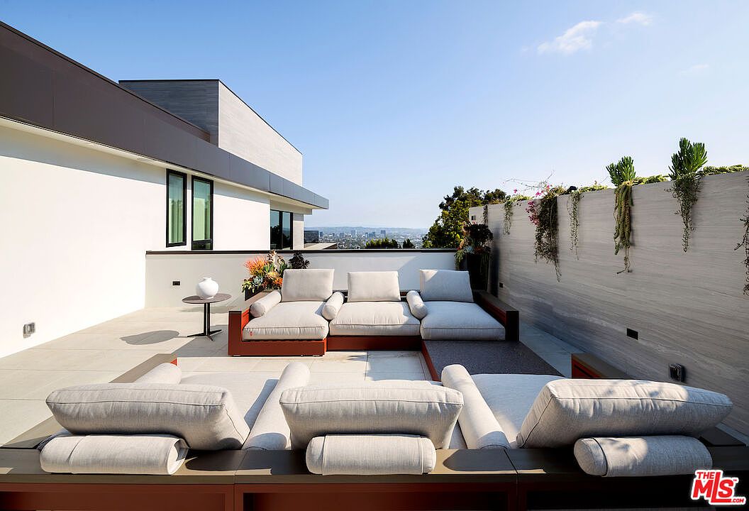 This expansive rooftop terrace features a modern, modular outdoor sectional sofa arranged in a U-shape, providing ample seating for relaxation. The space is defined by clean, minimalist lines, a light-toned stone wall with integrated greenery, and a panoramic view of the city skyline in the distance. The perspective is captured from a low angle, emphasizing the open-air layout and the seamless transition between the lounge area and the horizon.