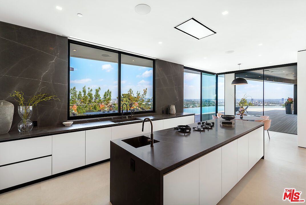 This sleek, modern kitchen features a large central island with a dark stone countertop and integrated gas cooktop, complemented by white cabinetry and dark stone wall cladding. Large glass sliding doors provide an expansive view of the outdoor deck and horizon, creating a seamless transition between indoor and outdoor living spaces. The minimalist design is enhanced by recessed lighting and a clean, open-concept layout that emphasizes the panoramic scenery.