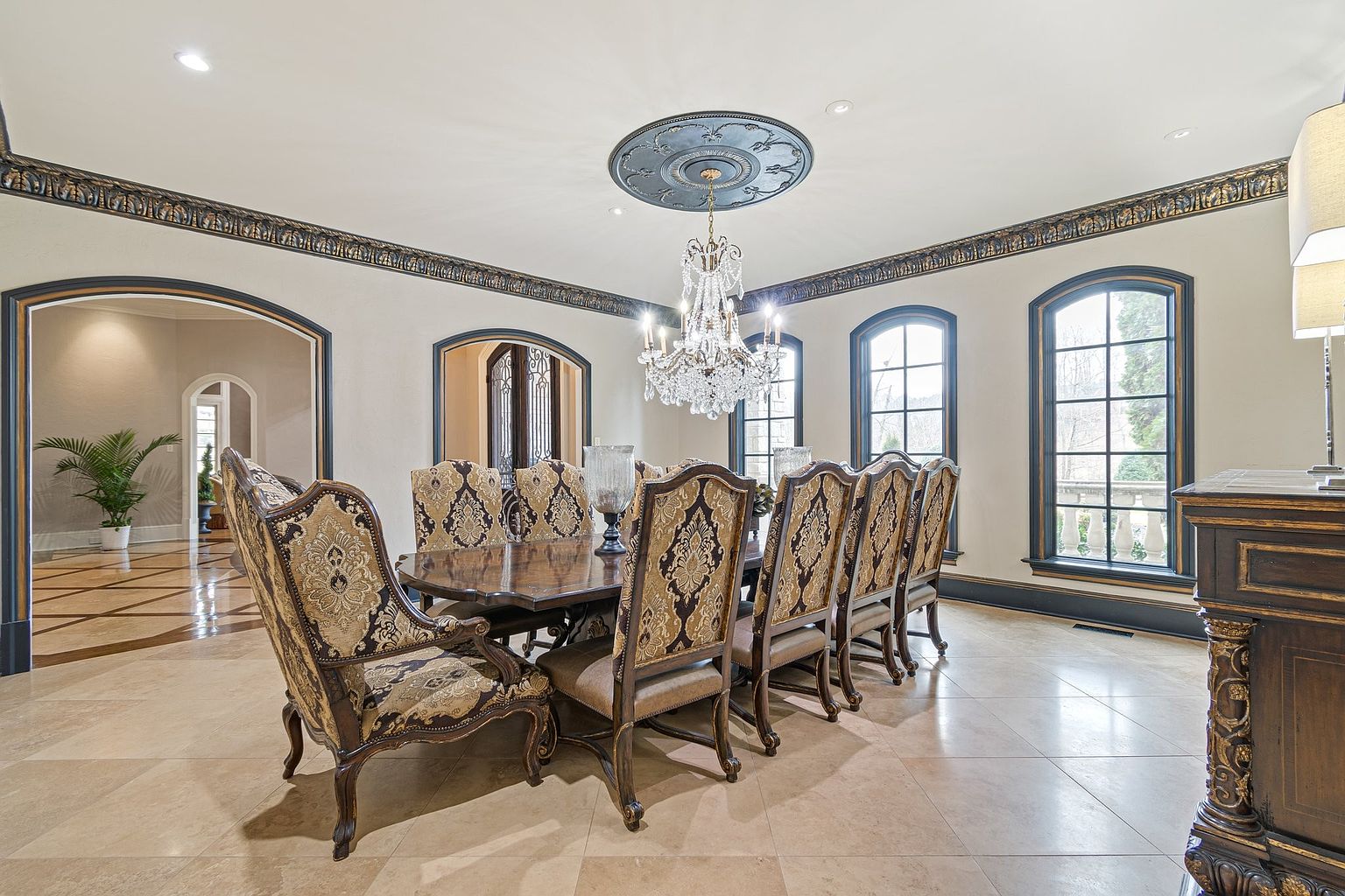 This is an interior shot of a formal dining room. The room features a large wooden dining table surrounded by ornate chairs with patterned upholstery. A crystal chandelier hangs above the table, and large windows provide natural light. The room is decorated with dark trim and molding, adding to the elegant and traditional style.