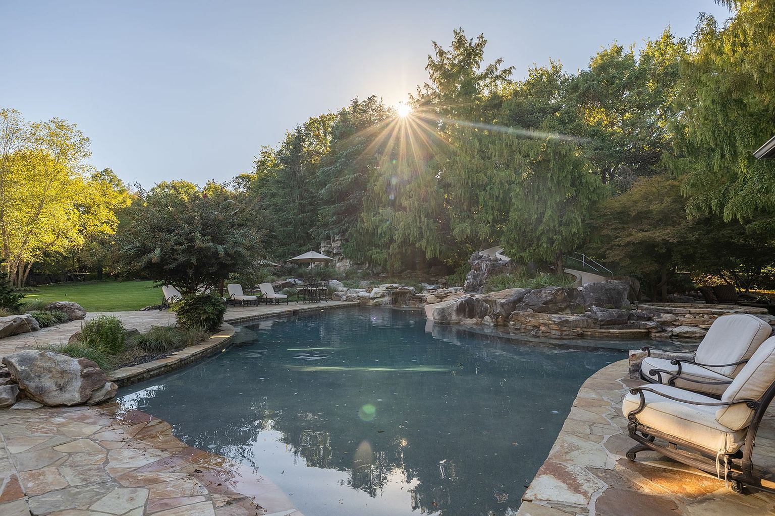 This image showcases a luxurious backyard pool area, complete with natural stone landscaping and mature trees providing shade and privacy. The pool features a rock waterfall element, and comfortable lounge chairs are positioned nearby for relaxation. The overall impression is one of a private oasis, perfect for outdoor entertaining and leisure.