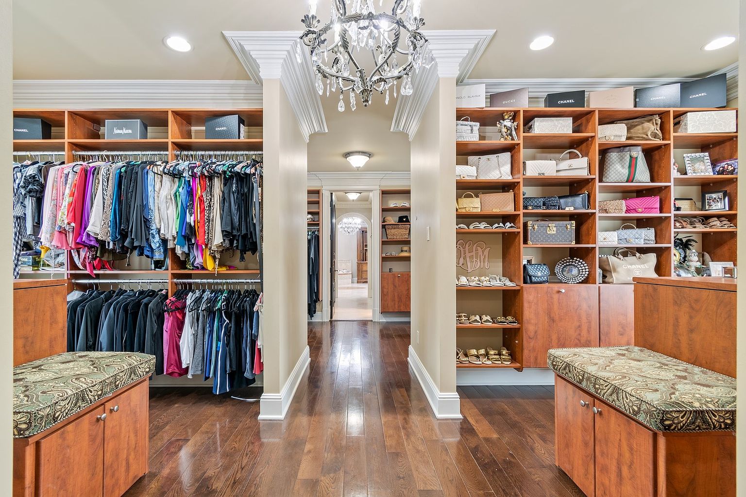 This is a luxurious walk-in closet featuring custom wood shelving and cabinetry. The closet is well-organized, showcasing clothing, handbags, and shoes. A decorative chandelier adds a touch of elegance, and the hardwood flooring contributes to the high-end feel of the space. The perspective is from the entrance of the closet, looking down the hallway.