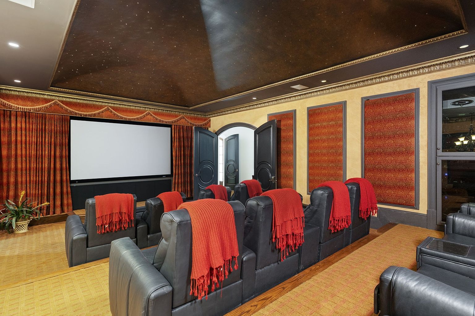 This is a home theater room featuring multiple rows of black leather recliners draped with red throws. A large screen is visible at the front, flanked by ornate curtains, and the ceiling is painted with a starry night effect. The room exudes a luxurious and comfortable atmosphere, perfect for entertainment.