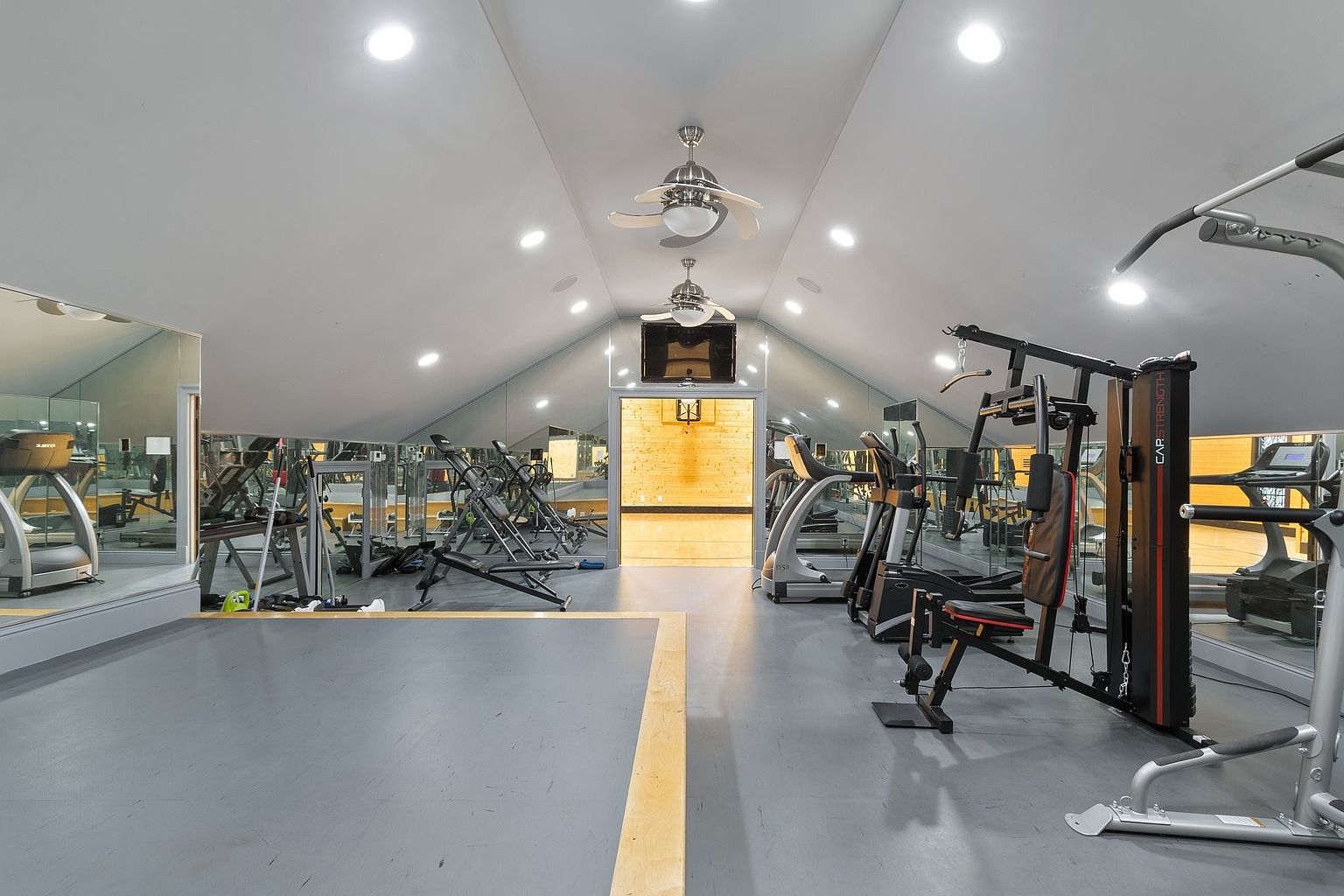 This is an interior shot of a well-equipped home gym. The room features a variety of exercise machines, including treadmills and weightlifting equipment, set against mirrored walls that enhance the sense of space. The room is well-lit with recessed lighting and ceiling fans, and a basketball hoop is visible at the end of the room.