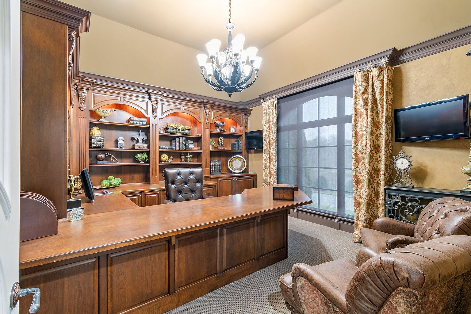This is an interior shot of a luxurious home office featuring a large, ornate wooden desk and built-in bookshelves filled with decorative items and books. Two leather armchairs sit near a large window with floral curtains, and a chandelier hangs from the ceiling, adding to the room's sophisticated and traditional style. A television is mounted on the wall.