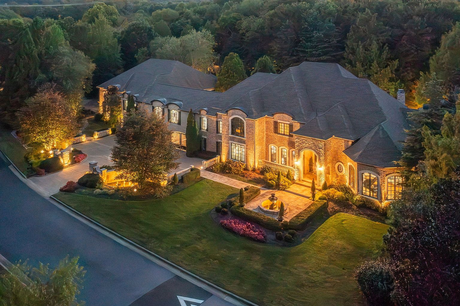 This aerial view showcases a grand estate with meticulously landscaped grounds, including manicured lawns, flower beds, and mature trees. The brick facade of the home is illuminated by exterior lighting, highlighting architectural details and creating a warm, inviting ambiance. A circular driveway and fountain add to the property's curb appeal and sense of luxury.