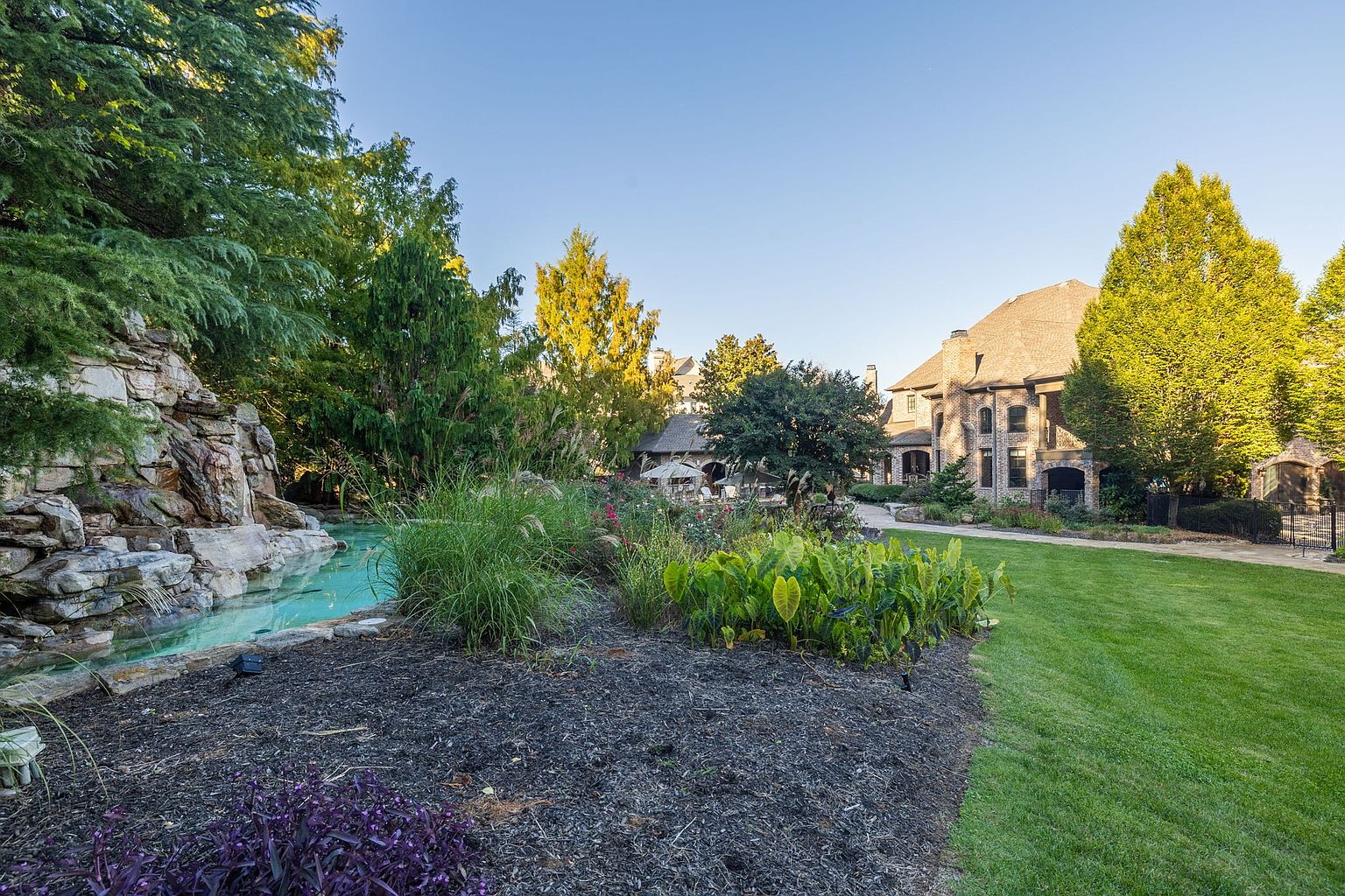 This image showcases a beautifully landscaped yard and garden area of a luxurious property. A rock waterfall feature adds a touch of elegance, flowing into a clear pool. The manicured lawn and meticulously arranged flower beds create a serene and inviting outdoor space, complemented by the grand architecture of the house in the background.