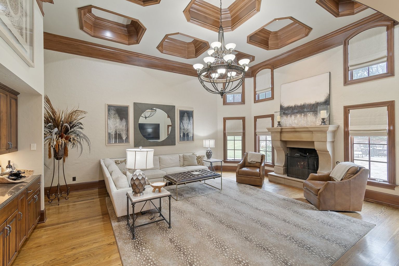 This is a spacious living room featuring hardwood floors, a large area rug, and a neutral-toned sectional sofa. The room is well-lit by a chandelier and natural light from the windows, which are framed by wooden trim. A stone fireplace and decorative artwork add to the room's elegance and charm, creating a warm and inviting atmosphere.