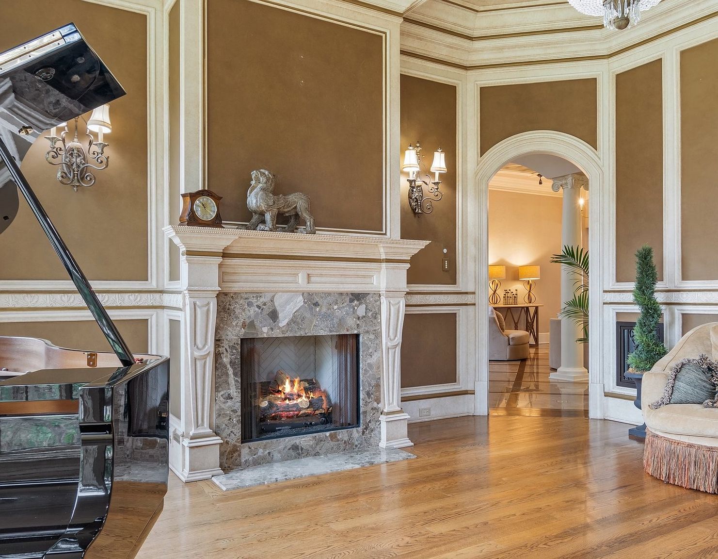 This is an elegant living room featuring a grand piano, a fireplace with a marble surround, and sophisticated wall paneling. The room is bathed in warm light, highlighting the hardwood floors and creating an inviting atmosphere. An arched doorway leads to another room, suggesting a spacious and well-designed home.