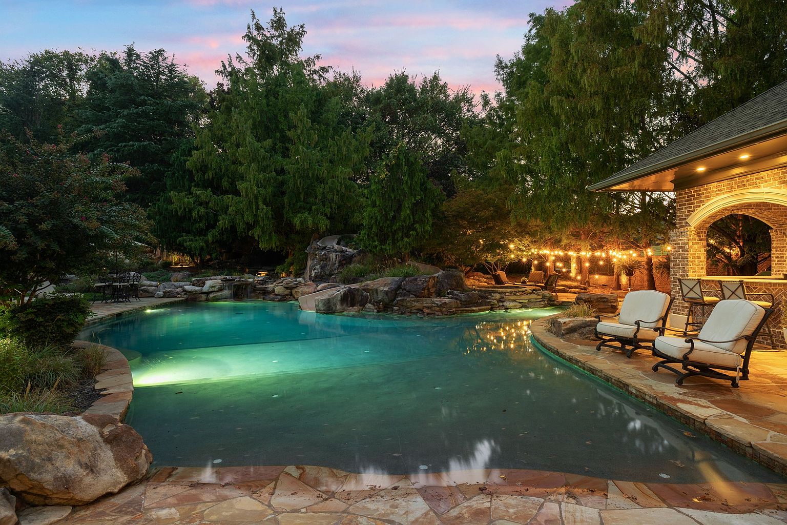 This image showcases a luxurious backyard pool and spa area at dusk. The pool features a natural rock waterfall and slide, with lush landscaping surrounding the perimeter. String lights add a warm ambiance to the outdoor bar area, complete with comfortable seating, creating an inviting and relaxing atmosphere.