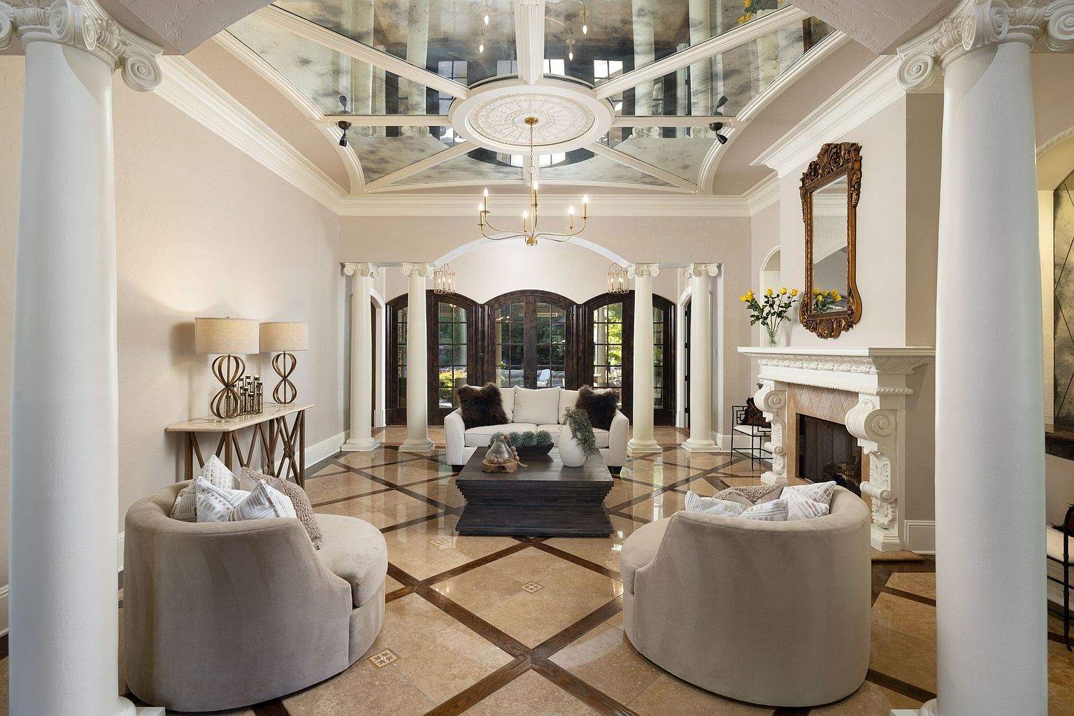 This is an elegant living room featuring a mirrored ceiling, ornate chandelier, and a fireplace with a decorative mirror above. The room is furnished with two round chairs, a white sofa, and a dark wood coffee table. The flooring is a patterned tile, and the space is framed by white columns, creating a luxurious and inviting atmosphere.