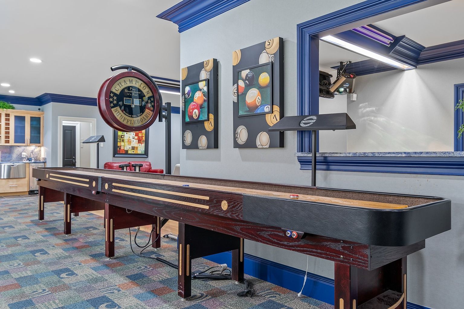This interior shot showcases a recreation room featuring a shuffleboard table as the central element. The room is decorated with billiard-themed artwork and has a pass-through window to another area. The overall impression is one of a well-appointed and entertaining space, perfect for leisure and relaxation.
