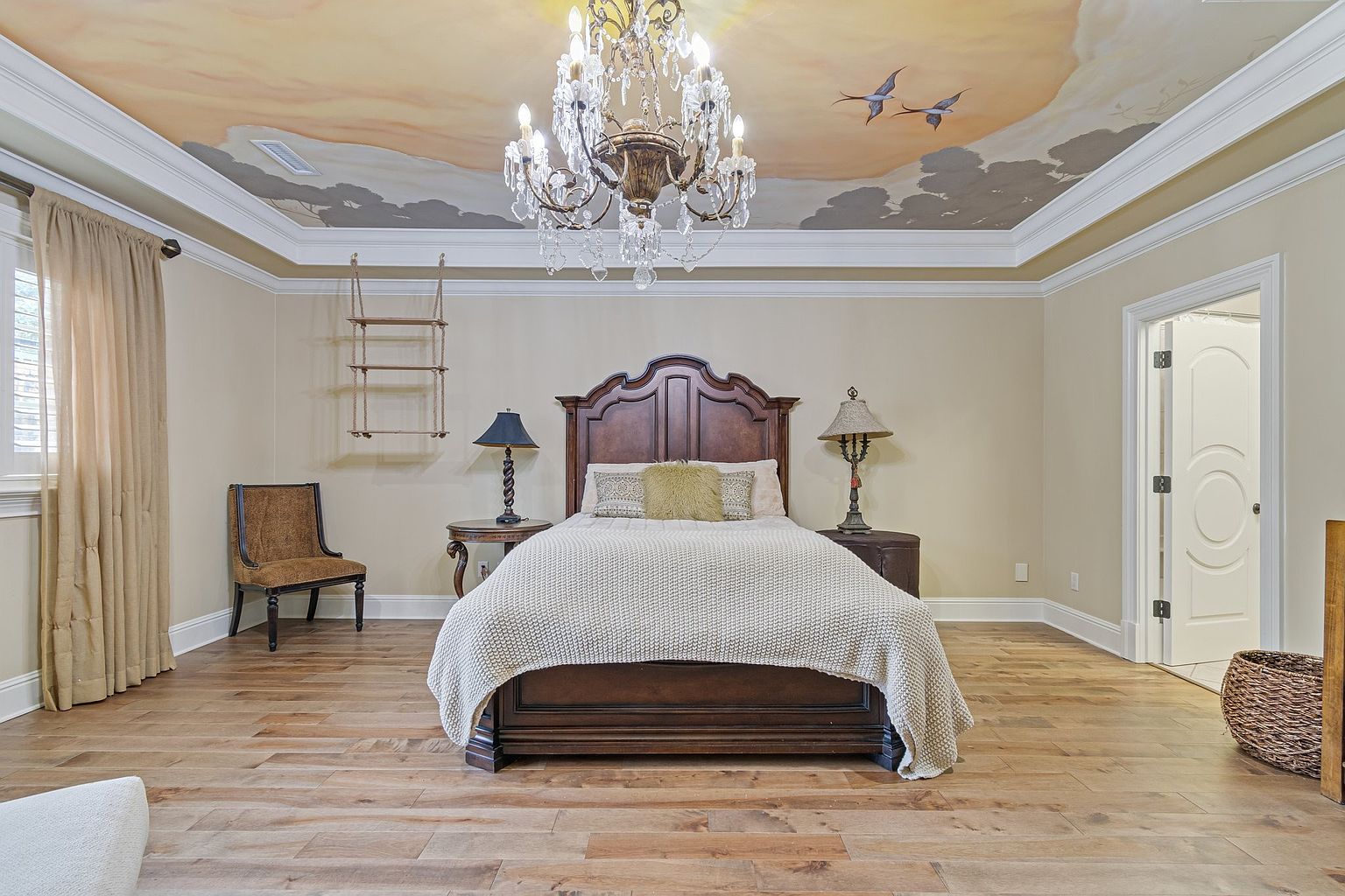 This is a primary bedroom featuring a wooden bed frame with a light-colored knitted blanket. The room has hardwood floors and a unique ceiling mural depicting a sky scene with birds. A large chandelier hangs above the bed, adding a touch of elegance.