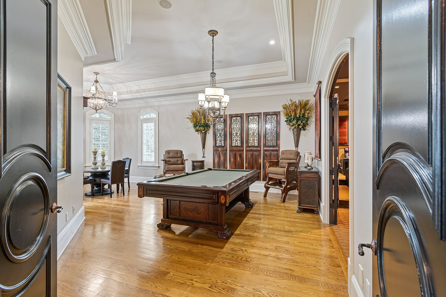 This is an interior shot of a luxurious living room featuring a billiards table as a focal point. The room is well-lit by chandeliers and natural light from arched windows, highlighting the hardwood floors and detailed ceiling trim. The space also includes seating areas and decorative elements, creating an elegant and inviting atmosphere.