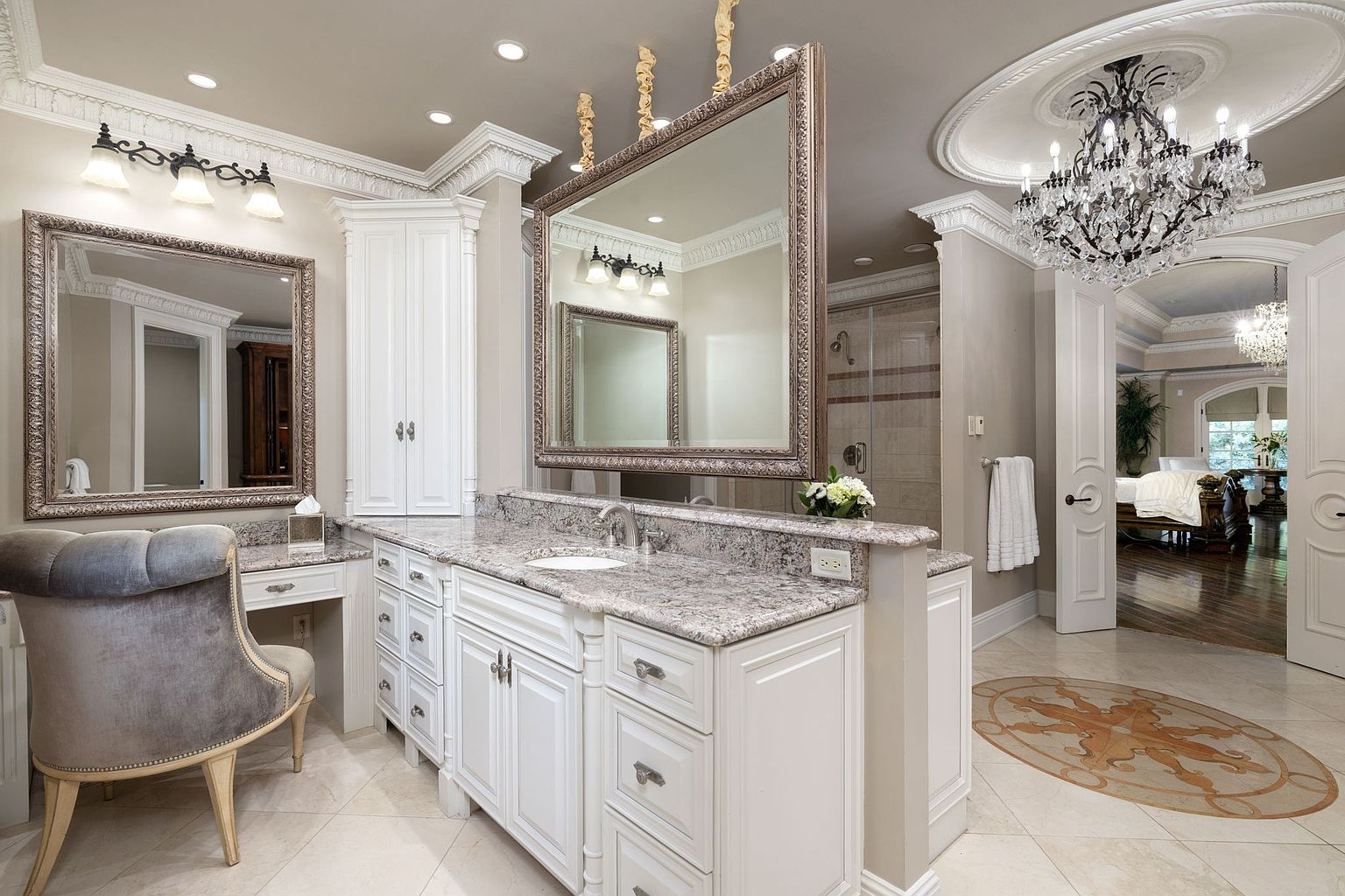 This is a luxurious primary bathroom featuring a double vanity with granite countertops and ornate mirrors. The cabinetry is white, and the room is illuminated by a chandelier and sconces. The bathroom opens into a bedroom area, showcasing a seamless transition and high-end finishes.