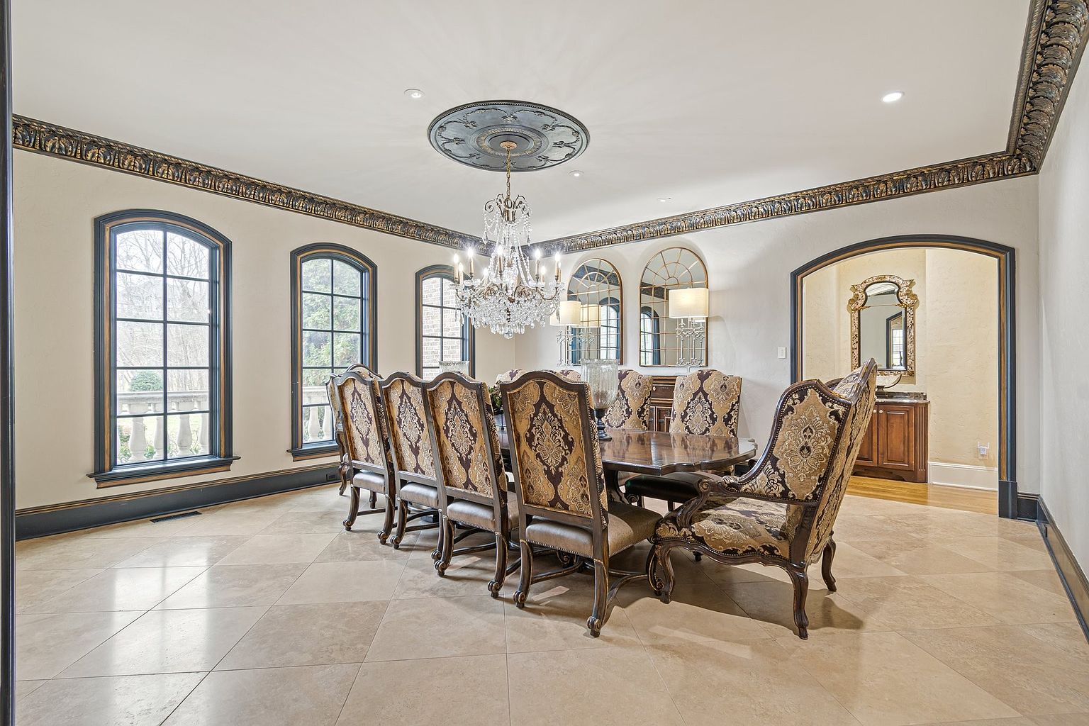 This is an interior shot of a formal dining room. The room features a large dining table with ornate chairs, a crystal chandelier, and arched windows. The walls are painted in a neutral tone with decorative molding, and the flooring is tiled. The overall impression is one of elegance and sophistication.