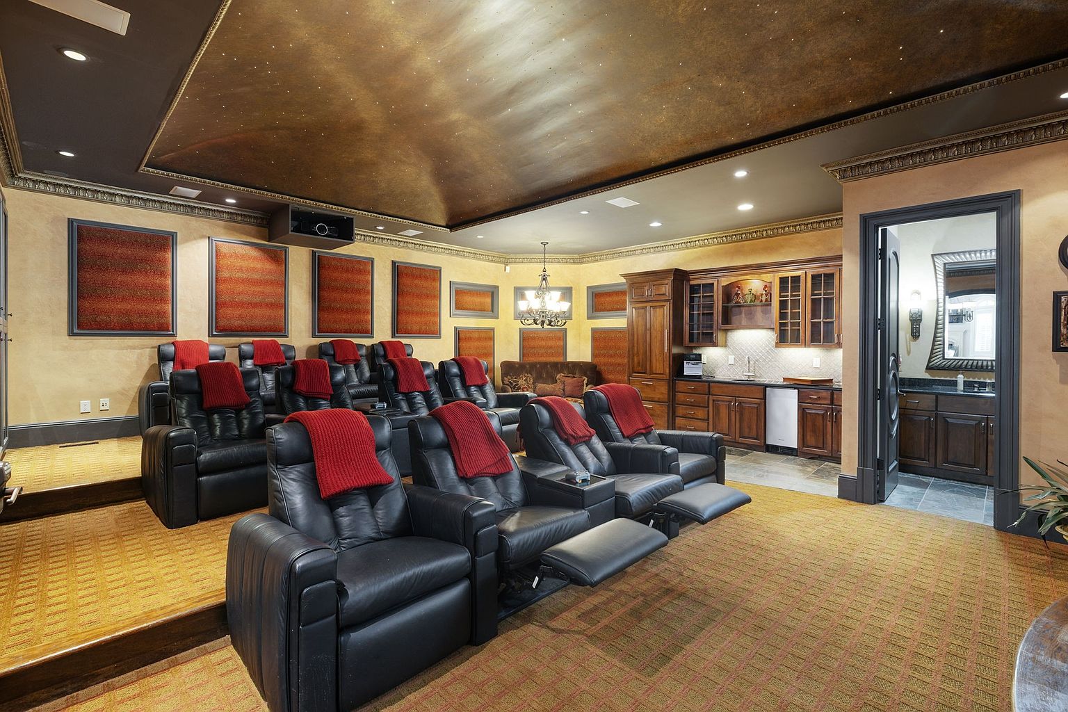 This is an interior shot of a home theater, featuring multiple rows of black leather recliners with red throws. The room includes a kitchenette area with wooden cabinets and a sink, as well as a doorway leading to a bathroom. The ceiling is decorated with a starry effect, and the walls are painted in warm tones, creating a luxurious and immersive viewing experience.