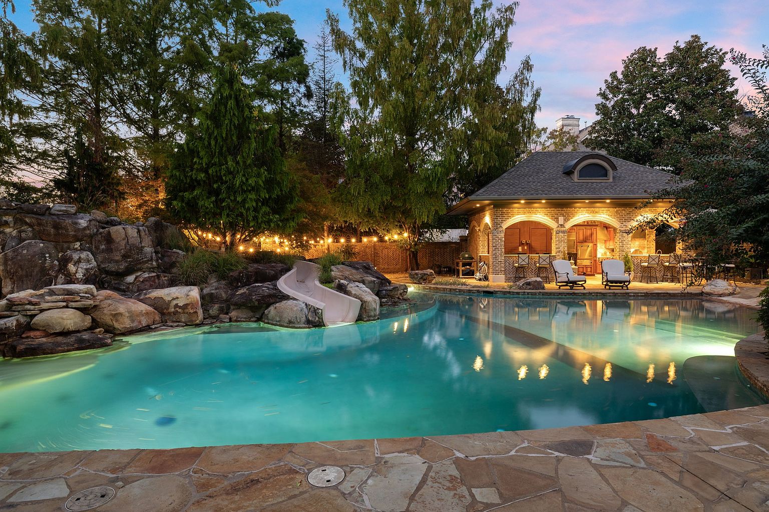 This image showcases a luxurious backyard pool area at dusk, featuring a custom pool with a slide built into a rock formation. A brick pool house with bar seating and lounge chairs sits adjacent to the pool, illuminated by warm lighting. The scene is framed by mature trees and string lights, creating an inviting and upscale outdoor living space.