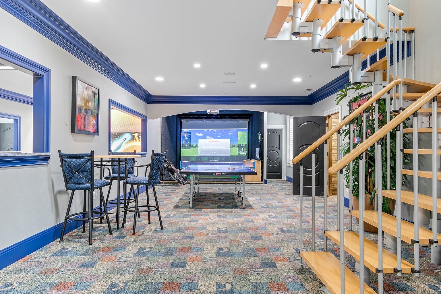 This is a spacious basement area featuring a golf simulator, ping pong table, and a bar area with stools. The room is decorated with blue trim and a patterned carpet. A modern staircase with wooden steps and metal railings leads to another level, adding a unique architectural element to the space.