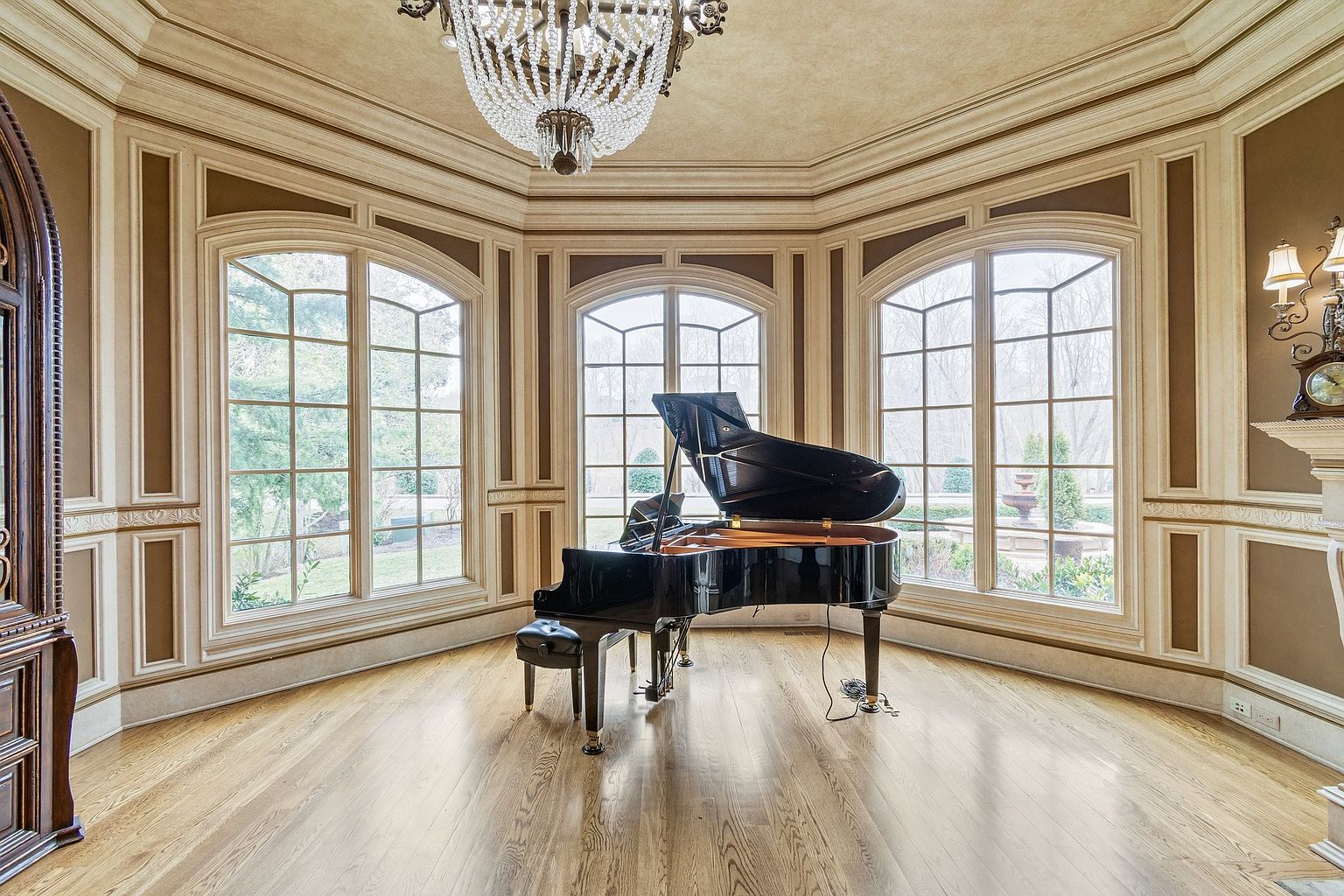 This grand living room features a black grand piano as its centerpiece, bathed in natural light from large arched windows overlooking a lush garden. The room is elegantly decorated with detailed wall paneling, a decorative chandelier, and hardwood floors, creating a luxurious and inviting atmosphere. The perspective is from the center of the room, capturing the piano and the windows.