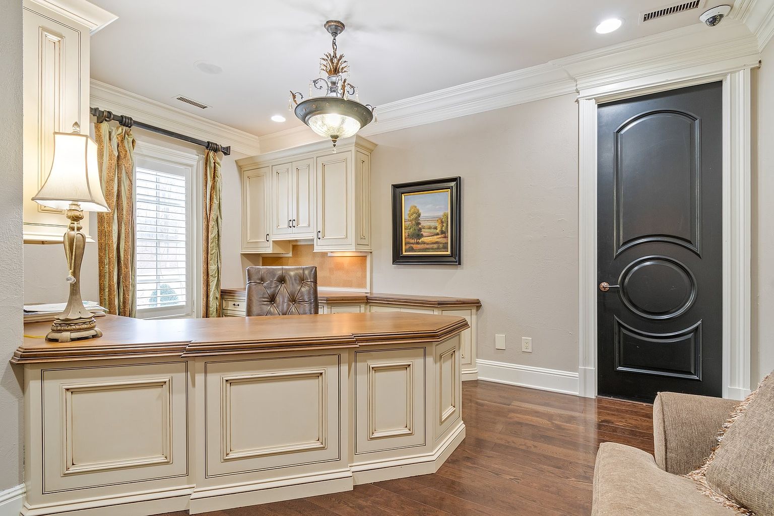 This is an interior shot of a home office or study. The room features a large, custom-built desk with cream-colored cabinetry and a wood top, complemented by overhead cabinets and a decorative chandelier. A black door with white trim is visible on the right, and a painting hangs on the wall, adding a touch of elegance to the space.