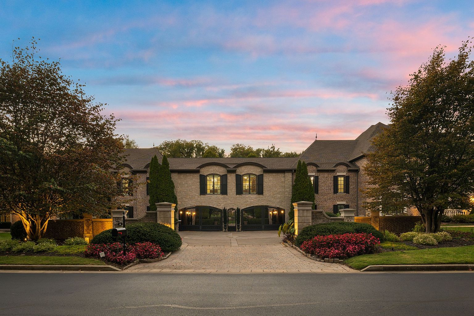 This is a front view of a grand, luxurious home featuring a stone facade with black shutters and arched windows above the garage doors. The driveway is paved with brick, leading to a porte-cochère flanked by manicured landscaping with red flowers and green shrubbery. The sky is painted with hues of pink and blue from the setting sun, adding to the overall elegance and curb appeal of the property.