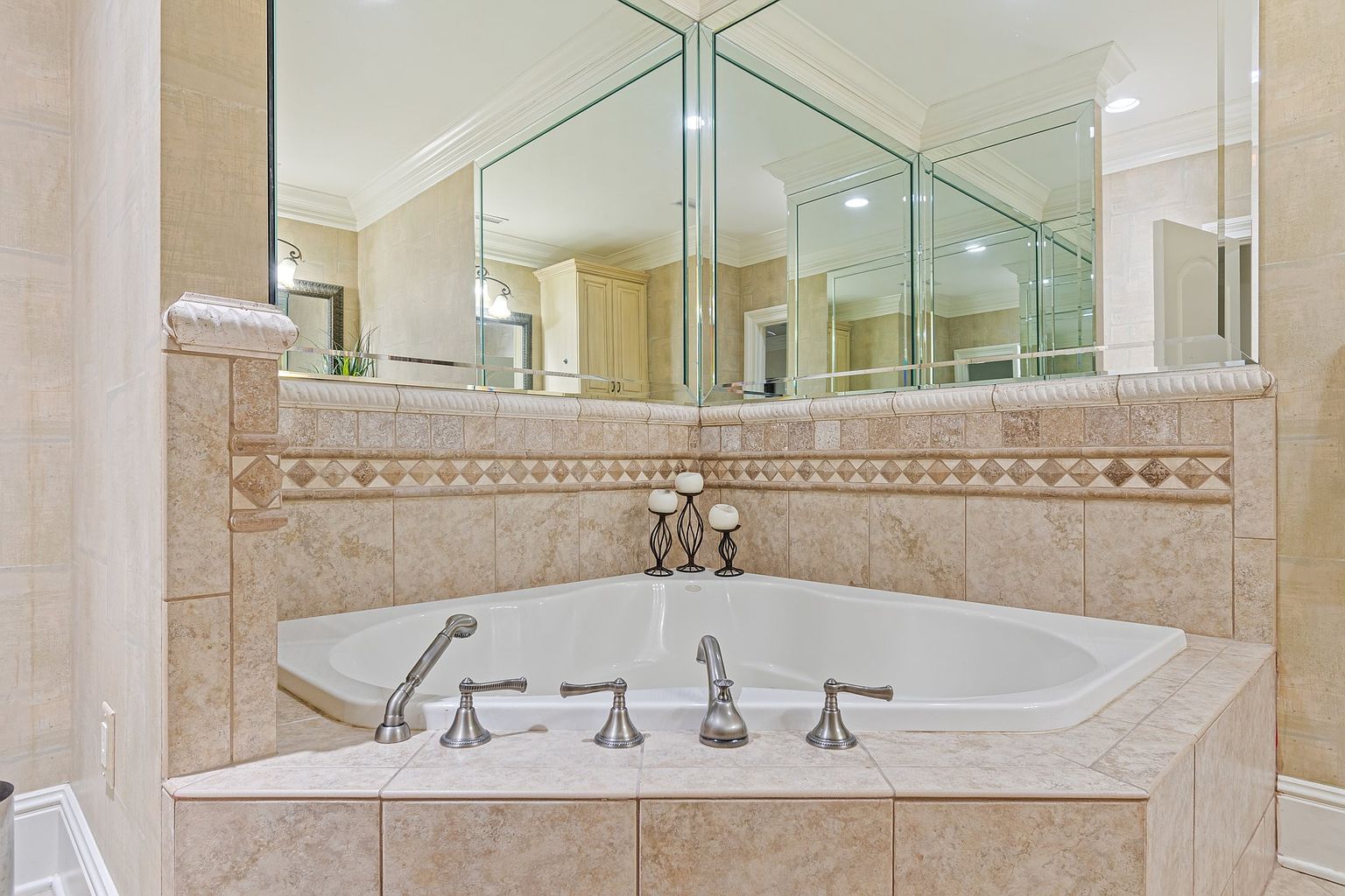 This is a close-up shot of a primary bathroom featuring a corner bathtub with a tiled surround. The tub has brushed nickel fixtures and is adorned with decorative candles. A large mirror reflects the bathroom's interior, creating a sense of spaciousness and luxury.