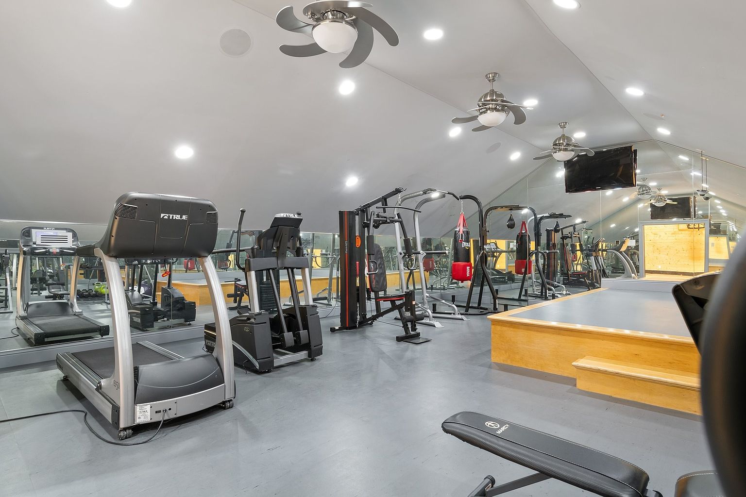 This is a spacious home gym featuring a variety of exercise equipment, including treadmills, elliptical machines, and weightlifting stations. The room has a mirrored wall, a raised platform, and multiple ceiling fans. The overall impression is a well-equipped and functional space for fitness activities.