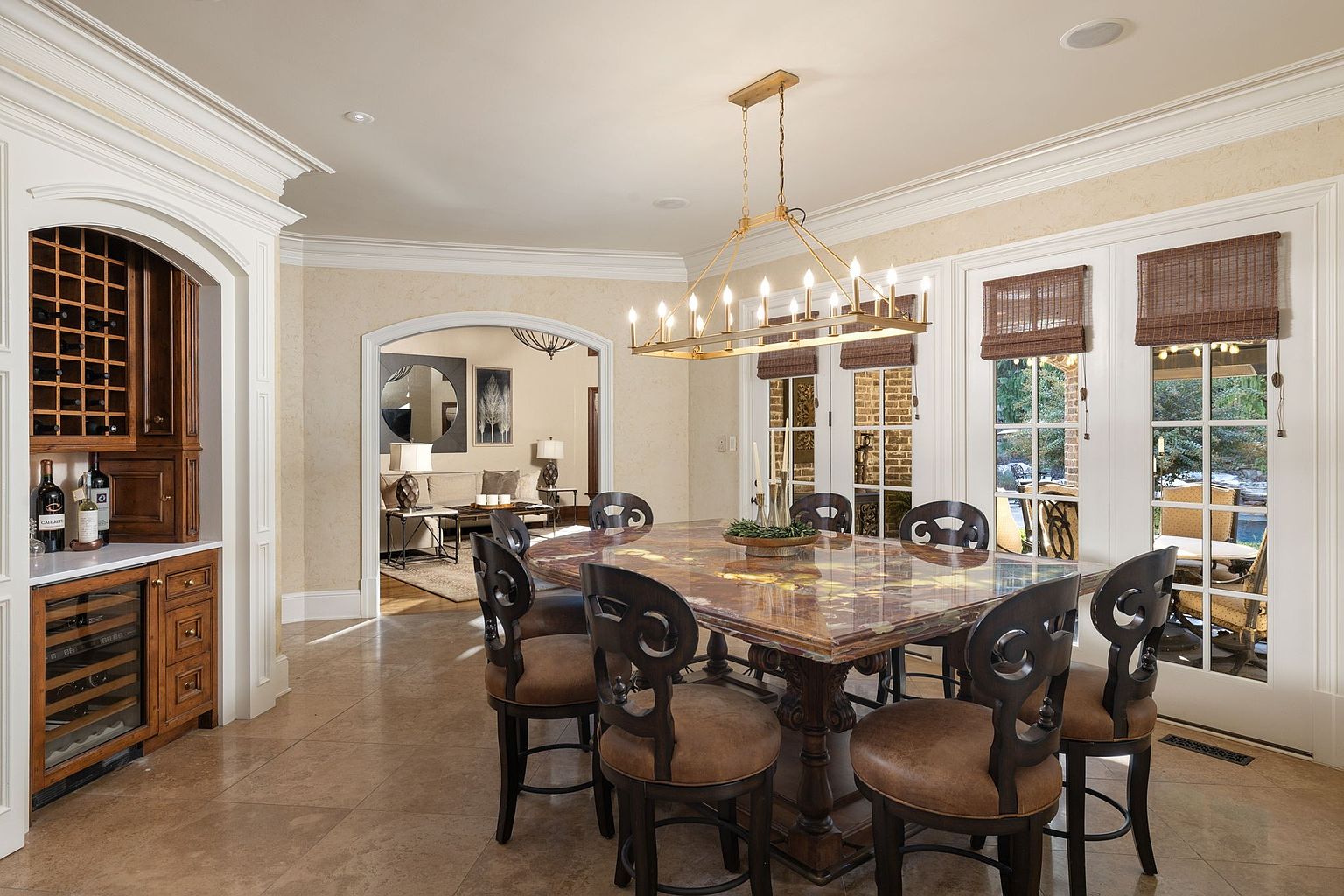 This is an interior shot of a dining room featuring a large, square dining table with a patterned top and ornate base, surrounded by six chairs with dark wood frames and brown leather seats. A rectangular chandelier hangs above the table, and natural light streams in through the French doors with woven shades, offering a glimpse of the outdoor patio. The room also includes a built-in wine storage unit and an arched doorway leading to another living space.