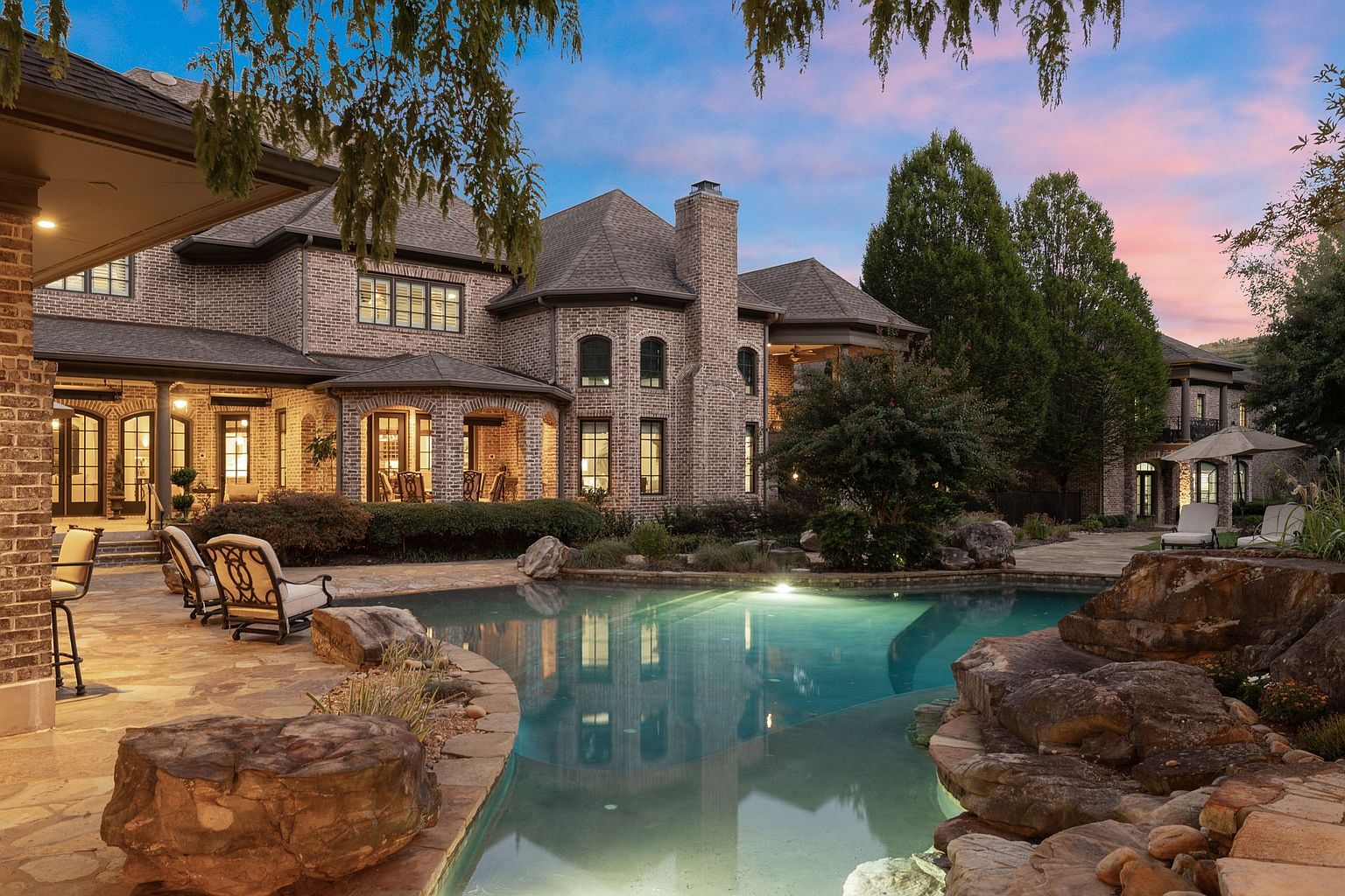 This is a stunning rear view of a large brick home featuring a luxurious pool and spa area. The pool is surrounded by natural rock formations and lush landscaping, creating a serene and private oasis. The architecture of the home is traditional with multiple levels and a prominent chimney, illuminated by warm lighting that reflects beautifully in the water.