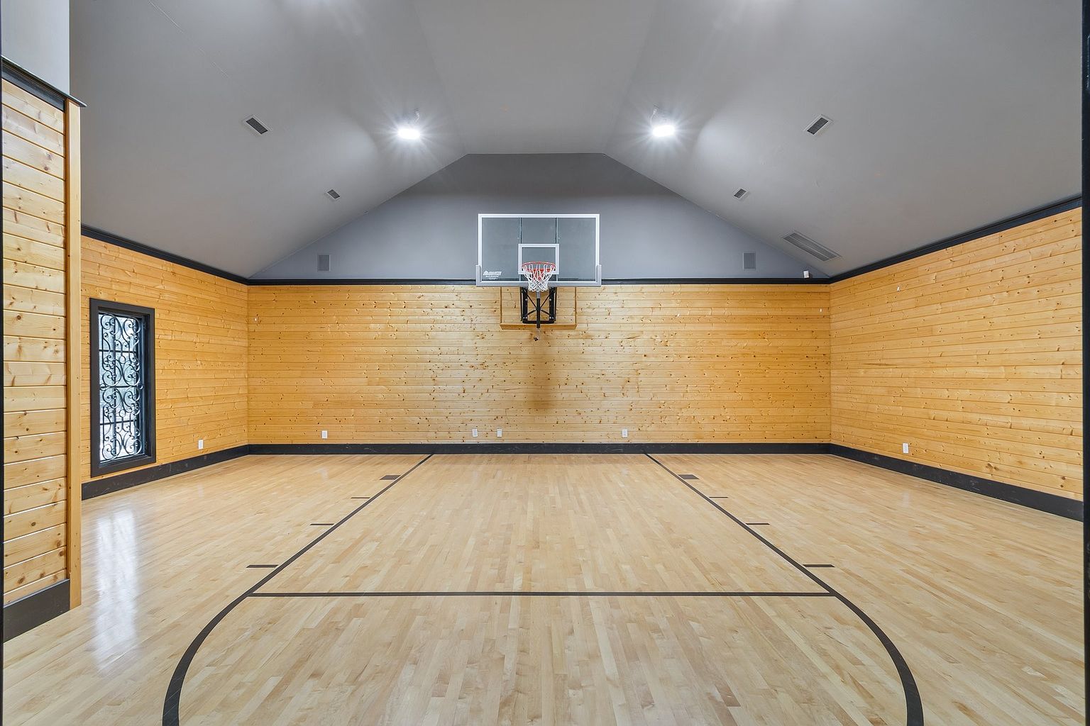 This is an interior shot of a home basketball court. The court features a wooden floor with black lines marking the boundaries, wood paneled walls, and a basketball hoop with a clear backboard. The room is well-lit with ceiling lights, and the overall impression is clean and spacious.