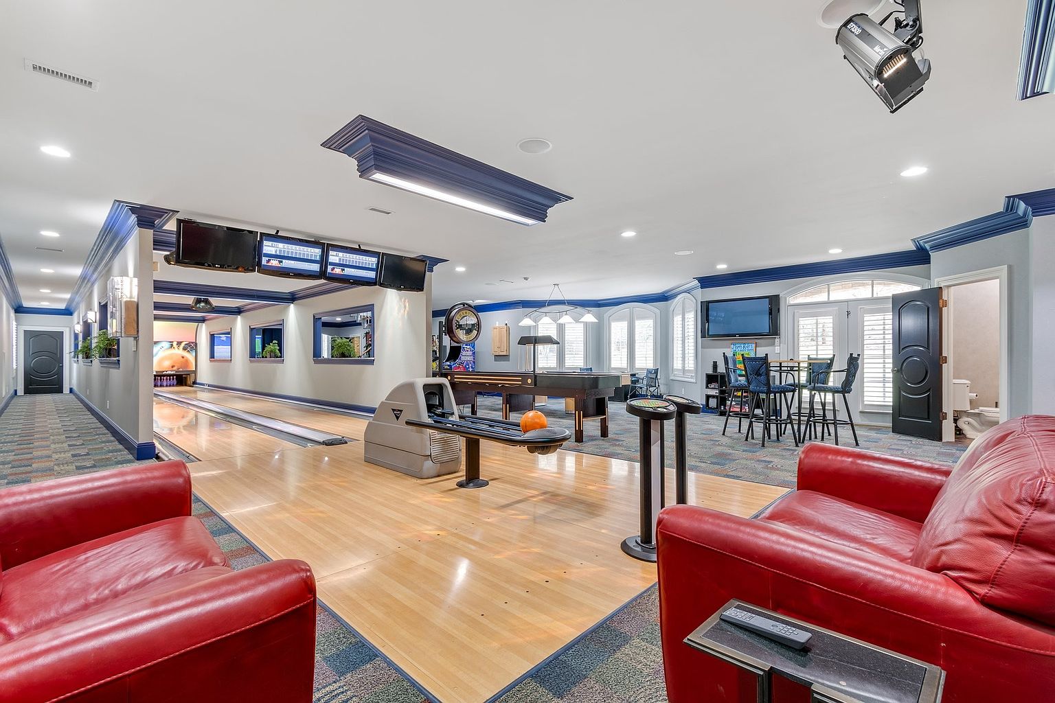 This is a spacious living room featuring a bowling alley and a pool table, creating an entertainment-focused space. The room is decorated with blue and white trim, hardwood floors, and comfortable red leather seating. Multiple televisions are mounted on the walls, enhancing the entertainment experience.