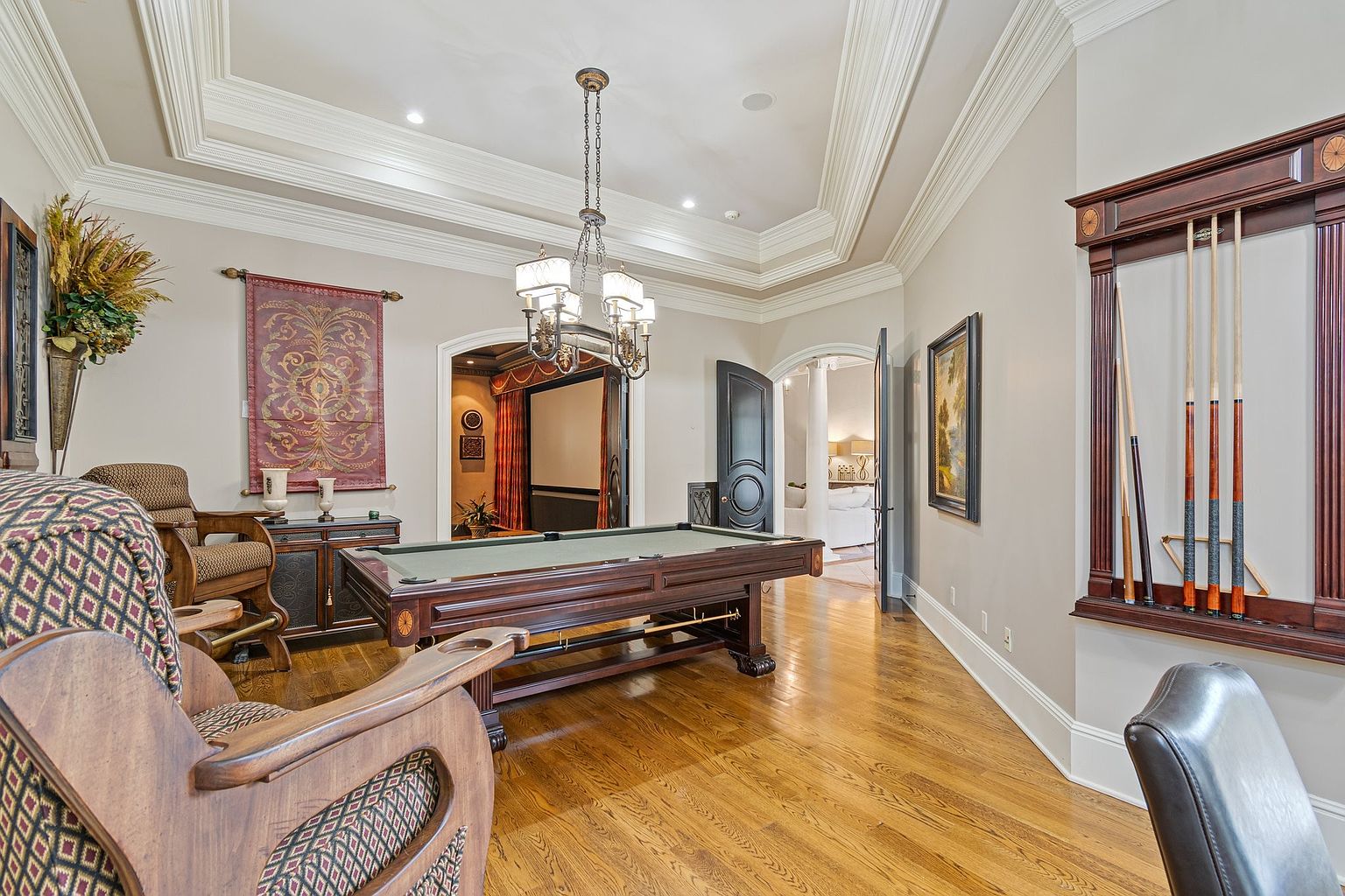 This interior shot showcases a luxurious living room featuring a billiard table as a central element. The room is adorned with elegant furnishings, including a patterned armchair and a decorative wall tapestry. The hardwood flooring and coffered ceiling add to the room's sophisticated ambiance, creating an inviting space for relaxation and entertainment.