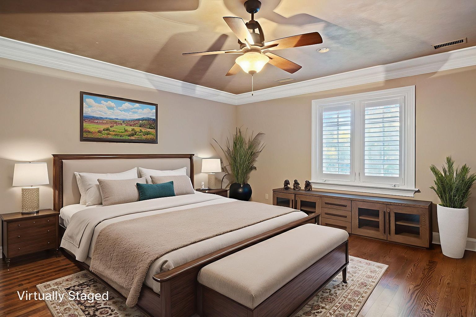 This is a staged primary bedroom featuring a large bed with a neutral-toned headboard and bedding, complemented by a dark wood frame. The room is decorated with a landscape painting, bedside tables with lamps, and a console table under a window with plantation shutters. A ceiling fan adds to the room's comfort, and the overall impression is one of calm and sophistication.